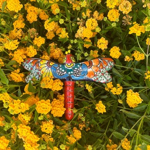 May include: A colorful ceramic dragonfly garden stake with a red body and intricately painted wings in blue, orange, and yellow. The dragonfly is set against a backdrop of vibrant yellow flowers and green foliage.