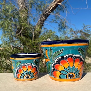 May include: Two ceramic flower pots with vibrant floral designs. The pots feature a teal background with orange, yellow, and blue floral patterns. The rims are dark blue with a yellow stripe. Suitable for indoor or outdoor use.