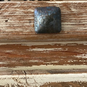 May include: Close-up of a square, hammered metal decorative nail head. The nail head is a dark, iridescent colour and is set against a distressed, rustic wooden surface with white paint.