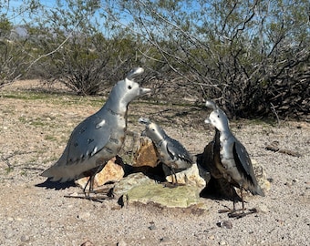 May include: Three metal sculptures of California quail standing on rocks. The sculptures are crafted from silver-toned sheet metal with visible welded seams and rivets, featuring the distinct topknot crests characteristic of the quail species.