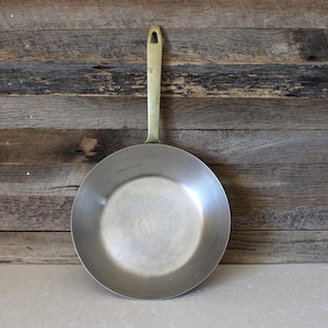 May include: A round metal frying pan with a smooth, light gray interior surface and a long, slender brass-colored handle featuring a triangular cutout at the end for hanging. The pan has a simple, functional design with a flat base and curved sides.