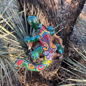 May include: A vibrant, hand-painted ceramic lizard sculpture. The lizard features a glossy green head and limbs, with a colorful body design including red, orange, yellow, and purple patterns. It is mounted on a piece of wood, set against a natural outdoor backdrop.