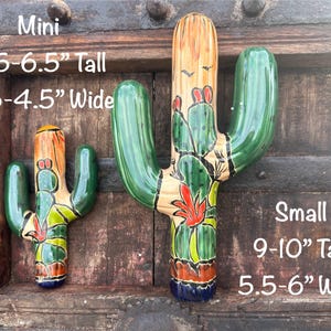 May include: Two ceramic cactus sculptures, one small and one mini, with hand-painted details. The mini cactus is 5.5-6.5 inches tall and 3.5-4.5 inches wide. The larger cactus is 9-10 inches tall and 5.5-6 inches wide. Both have green, orange, and red accents.
