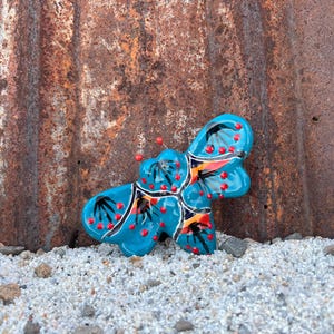 May include: A bright turquoise ceramic butterfly-shaped object featuring black painted leaf-like patterns and small raised red dots across its surface. The piece has a glossy finish and is shaped with multiple rounded wings and a central body section.