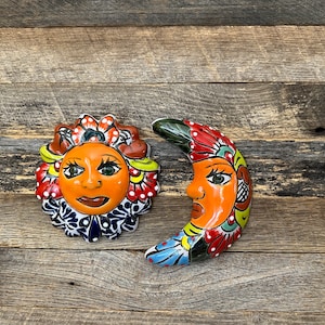 May include: A pair of colorful ceramic wall hangings depicting a sun and a crescent moon. The sun has a smiling face with green eyes and a red mouth. The moon has a frowning face with green eyes and a red mouth. Both are decorated with floral patterns in shades of red, yellow, blue, and green.