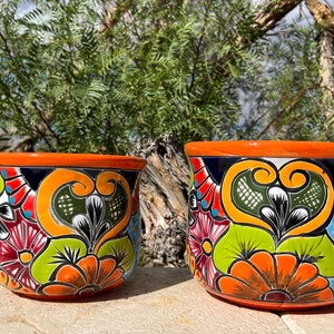 May include: Two hand-painted ceramic flower pots. The pots have an orange rim and are decorated with colorful floral patterns in shades of orange, green, blue, and red. The pots are sitting on a stone surface.