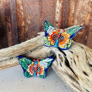 May include: Two ceramic butterfly figurines with vibrant colors. The butterflies have blue wings with green and orange details, and a central flower design. They rest on a piece of driftwood against a rusty metal background.