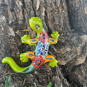 May include: A green ceramic lizard with colourful floral designs. The lizard is painted with red, blue, yellow, and orange flowers. The lizard is perched on a tree trunk.