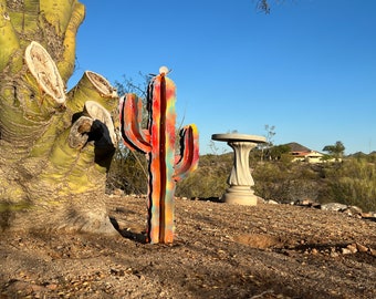May include: A colorful metal cactus sculpture with a red, orange, yellow, blue, and green paint design. The sculpture is standing on a dirt surface in front of a tree.