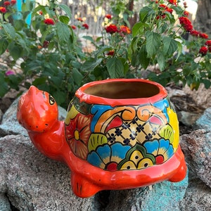 May include: A brightly coloured ceramic turtle planter. The turtle is predominantly orange with a shell decorated with floral and geometric patterns in blue, yellow, and green. The planter is set against a backdrop of rocks and greenery.