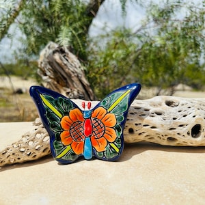 May include: A blue and green ceramic butterfly with a yellow and orange flower design. The butterfly is painted in a traditional Mexican style.