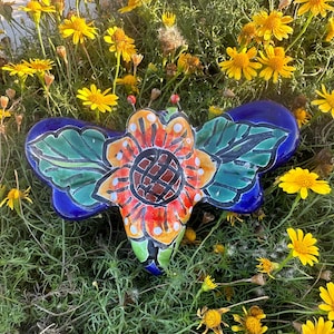 May include: A colorful ceramic butterfly-shaped garden ornament. The wings are a deep blue, and the body features a flower design with orange, red, and green accents. The ornament is surrounded by yellow flowers and green foliage.