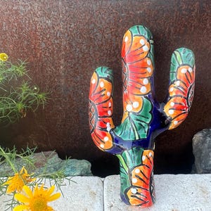Könnte beinhalten: Eine farbenfrohe Keramik-Kaktus-Skulptur mit einem lebendigen Design. Der Kaktus hat orange, rote, grüne und weiße Blumenmuster auf einem blauen Sockel. Ein einzigartiger Dekorationsartikel für das Zuhause.