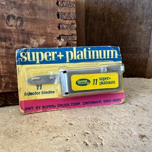 May include: A vintage package of Super+ Platinum injector blades. The package features a yellow and pink color scheme with the text "super+platinum" and "11 injector blades". The blades are in a silver metal holder.