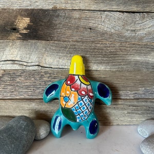 May include: A ceramic turtle figurine with a turquoise body, yellow head, and colourful details. The turtle's shell features red, orange, and white patterns. The figurine is displayed against a rustic wooden background.