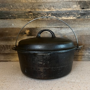 May include: A black cast iron Dutch oven with a lid and a metal handle. The pot is round with a slightly textured surface. The lid has a handle on top. The background is a wooden wall.