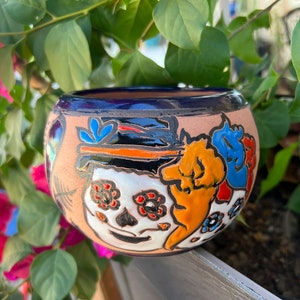 May include: A round, hand-painted ceramic pot with a dark blue rim and a tan body. The pot features a colorful Day of the Dead sugar skull design with orange, blue, and red accents. The pot is approximately 4 inches in diameter.