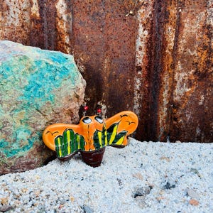 May include: A whimsical, orange ceramic butterfly figurine with black, yellow, and green details. The butterfly has large eyes and red antennae. It is set against a backdrop of a rusty metal sheet and a turquoise rock, resting on a bed of small white pebbles.