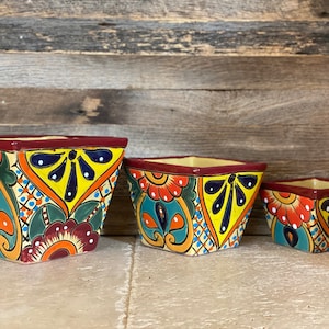 May include: A trio of square ceramic planters with a vibrant, hand-painted design. Each planter has a burgundy rim, yellow interior, and detailed patterns in orange, teal, and blue. The sizes vary, creating a tiered display.