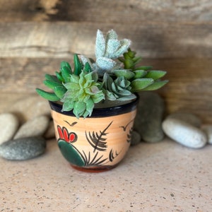Peut inclure: Petit pot en céramique peint à la main, base beige, bord noir et motifs floraux colorés. Il est rempli de diverses succulentes artificielles vertes et grises. Des pierres décoratives entourent le pot.