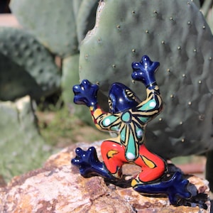 May include: A colorful ceramic frog figurine with blue, red, yellow, and green accents. The frog is posed on a rock with its arms raised, set against a backdrop of a green cactus. A decorative item.