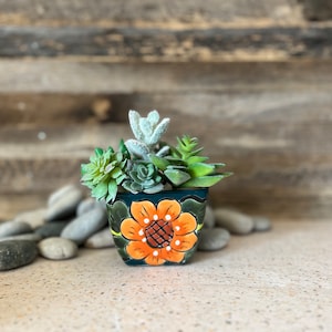 May include: A small, hand-painted terracotta pot with a green and orange floral design. The pot is filled with a variety of succulents.