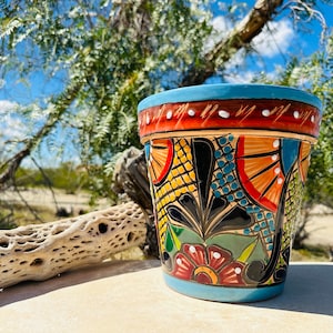 May include: A colorful ceramic planter with a blue base and intricate floral designs in shades of red, orange, yellow, green, and black. The planter is sitting on a light brown surface.