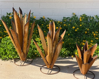May include: Three metal agave plant sculptures of varying heights, each featuring pointed, rust-colored leaves arranged in a rosette pattern. Each sculpture is supported by a circular metal base, creating a tiered display of desert-inspired art.