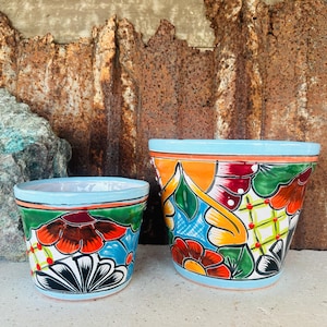 May include: Two hand-painted ceramic flower pots. The pots feature a light blue rim and base, with vibrant floral designs in orange, red, green, and blue. The larger pot is approximately 8 inches in diameter, and the smaller pot is approximately 6 inches in diameter.