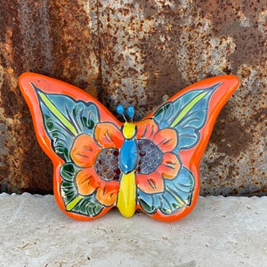 May include: A hand-painted ceramic butterfly wall decor with orange, yellow, green, blue, and brown accents. The butterfly has a floral design and is perfect for adding a touch of whimsy to any home.