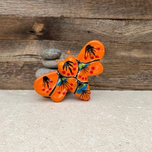 May include: A ceramic butterfly figurine in bright orange with black and red details. The wings have blue and green accents. The butterfly is resting on a light-colored surface, with a stack of gray stones and a wooden background.