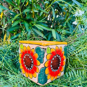 May include: A square terracotta planter with a hand-painted design of sunflowers in shades of orange, yellow, red, and green. The planter has a light brown background and a yellow rim.