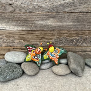 May include: Two colorful ceramic butterflies with green, yellow, orange, red, and white paint. The butterflies are sitting on a pile of gray stones.