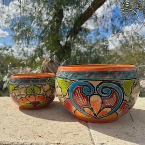 May include: Two hand-painted ceramic bowls with vibrant floral and geometric designs. The bowls feature a mix of colours, including orange, green, blue, and red, set against a cream background. The larger bowl is approximately 25 cm in diameter, while the smaller one is about 15 cm.