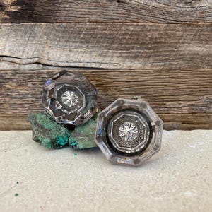 May include: Two clear, faceted glass door knobs with a starburst design, resting on a piece of green stone. The knobs have an octagonal shape and are set against a weathered wood background.