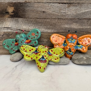 May include: Three ceramic butterfly figurines in shades of green, orange, and yellow, with red and blue accents. Each butterfly has antennae and is resting on a bed of gray stones. The background is a rustic wooden surface.