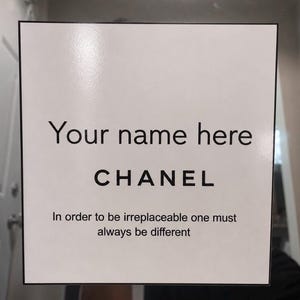 May include: A white square sign with black text that reads "Your name here" above "CHANEL". Below, the sign states "In order to be irreplaceable one must always be different". The sign has a black border.