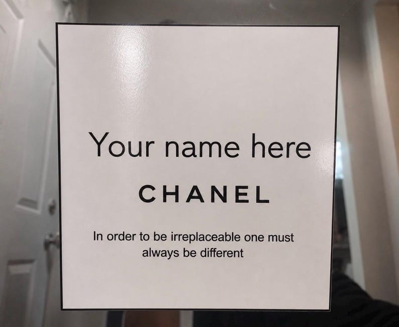 May include: A white square sign with black text that reads "Your name here" above "CHANEL". Below, the sign states "In order to be irreplaceable one must always be different". The sign has a black border.
