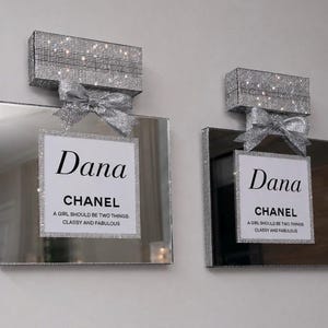 May include: Two decorative perfume bottle-shaped wall art pieces. Each features a mirrored surface, a sparkling crystal-covered top, and a silver bow. The center of each piece has a white label with the name "Dana" and the word "CHANEL". The text reads "A GIRL SHOULD BE TWO THINGS: CLASSY AND FABULOUS."