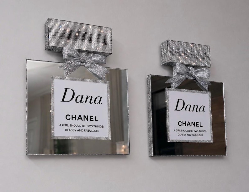 May include: Two decorative perfume bottle-shaped wall art pieces. Each features a mirrored surface, a sparkling crystal-covered top, and a silver bow. The center of each piece has a white label with the name "Dana" and the word "CHANEL". The text reads "A GIRL SHOULD BE TWO THINGS: CLASSY AND FABULOUS."