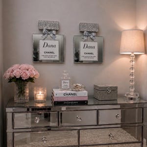 May include: A mirrored dresser with crystal drawer pulls and a mirrored top. Two decorative perfume bottle wall hangings with glitter tops and bows are above the dresser. A lamp, flowers, books, and a jewelry box are on the dresser.