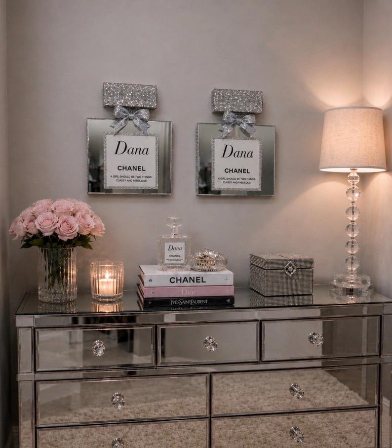 May include: A mirrored dresser with crystal drawer pulls and a mirrored top. Two decorative perfume bottle wall hangings with glitter tops and bows are above the dresser. A lamp, flowers, books, and a jewelry box are on the dresser.