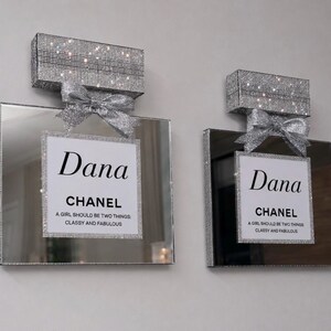 May include: Two decorative perfume bottle-shaped wall art pieces. Each features a mirrored surface, a sparkling silver bow, and a rectangular top covered in shimmering crystals. The center label reads "Dana CHANEL" with a quote.