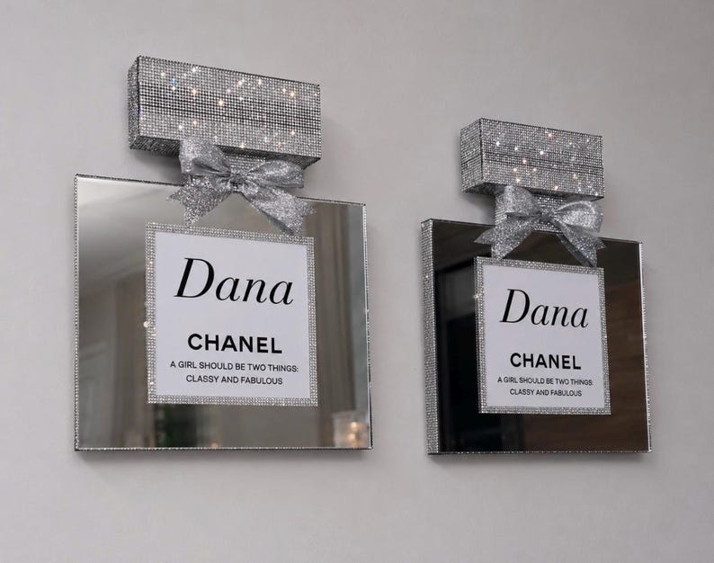 May include: Two decorative perfume bottle-shaped wall art pieces. Each features a mirrored surface, a sparkling silver bow, and a rectangular top covered in shimmering crystals. The center label reads "Dana CHANEL" with a quote.