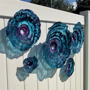 May include: A collection of five circular wall art pieces featuring a swirling pattern of deep blue, turquoise, and purple hues. Each piece has a concave centre and a textured, organic edge, creating a layered appearance with varying sizes.
