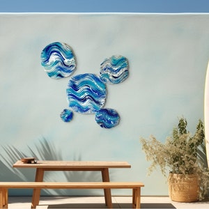 May include: Abstract wall art in blue and white, mimicking waves, adorns a light blue wall. A wooden table and bench are positioned below, with a surfboard nearby. The artwork and setting create a coastal atmosphere, reminiscent of a seaside location.