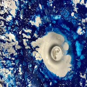 May include: A decorative glass bowl featuring a vibrant abstract pattern of deep royal blue and metallic silver. The design displays a swirling, marbled effect with concentrated silver pigment at the center and scattered silver flecks throughout.