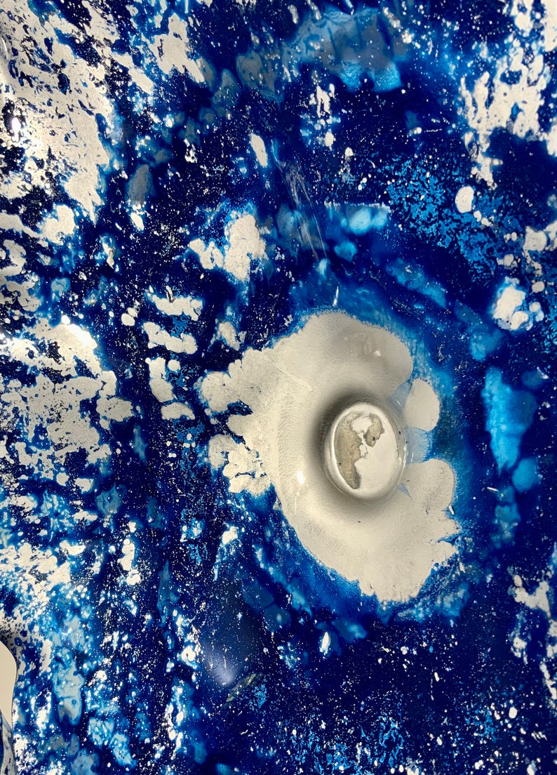 May include: A decorative glass bowl featuring a vibrant abstract pattern of deep royal blue and metallic silver. The design displays a swirling, marbled effect with concentrated silver pigment at the center and scattered silver flecks throughout.