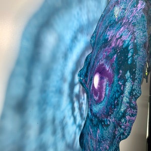 May include: A wall-mounted glass sculpture featuring a wavy, organic edge with a deep purple centre that transitions into swirling shades of blue and teal. The translucent material creates a soft, diffused blue shadow on the wall behind it.