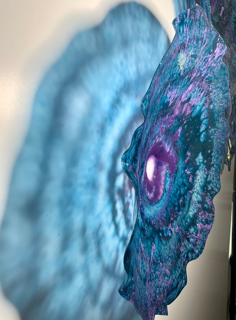 May include: A wall-mounted glass sculpture featuring a wavy, organic edge with a deep purple centre that transitions into swirling shades of blue and teal. The translucent material creates a soft, diffused blue shadow on the wall behind it.